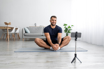 Happy european millennial guy sitting on mat looking at smartphone webcam recording sports blog in...