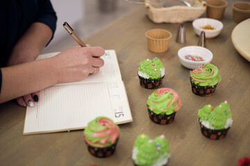 unrecognizable woman writing down notes while making cupcakes at cozy kitchen. cooking as a hobby, a master class for a pastry chef