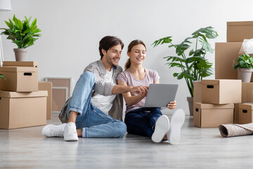 Smiling caucasian young husband and wife look at pc among cardboard boxes with stuff in room