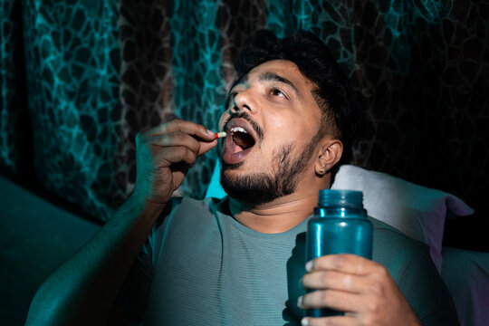 Young man taking medicine pills or tablets on bed while sleeping at night - conept of insomnia, unhealthy lifestyle, depression and illness.