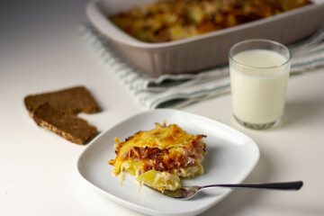 A piece of potato casserole on a square plate with a fork. Next to the casserole pan, a glass of milk, pieces of rye bread