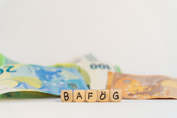 German word for student loans, BAFÖG, spelled with wooden letters wooden cube on a plain white background with banknotes, concept image 