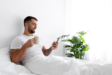 Smiling millennial muscular european man drink coffee, chatting on smartphone and enjoy good morning