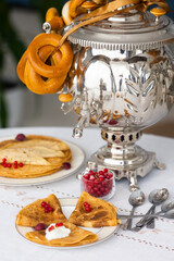 Russian traditional pancakes or blini with sour cream and red currant berries on table laid with samovar with bagels for Maslenitsa holiday or carnival   