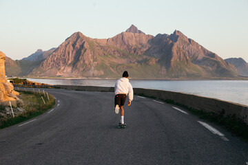 Millennial hipster skateboard away into sunset on summer evening. Young man in hoodie and shorts ride longboard in epic landscape scenery. Outdoor lifestyle vibes