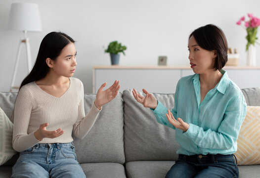Unhappy mature Asian woman and her adult daughter having fight, arguing with each other on couch in living room