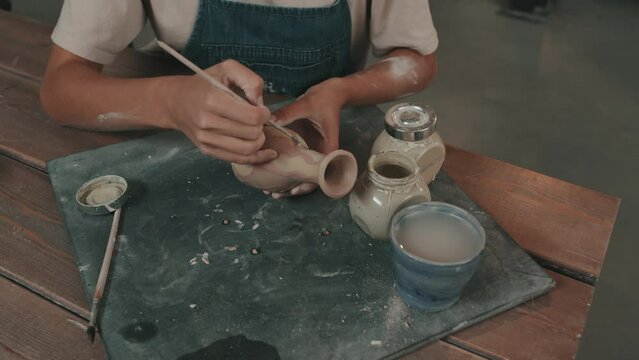 High-angle of cropped female artist with dry clay on hands wearing apron, sitting at table in modern studio, coloring handmade ceramic vase with brush and paint
