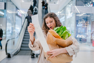 A woman in shock looks into a paper check from a supermarket in a shopping center against the background of an escalator and holds a package with fresh products, price increase - obrazy, fototapety, plakaty