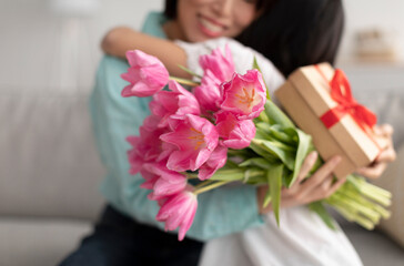 Unrecognizable Asian girl hugging granny, giving her tulips and gift for Women's Day at home, selective focus on flowers