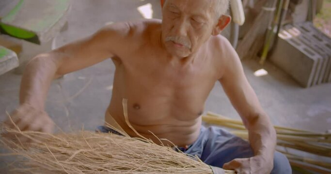 Asian male farmer who is an elderly person makes brooms from the grass that has many names called Paddy's lucerne, Queensland hemp , Arrowleaf sida , Common sida , Cuba juite for extra income.