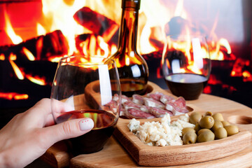 Female hand hold wine glass against wine table with appetizers for wine and electric fireplace. Manicure with red hearts. Romantic dinner at home concept.