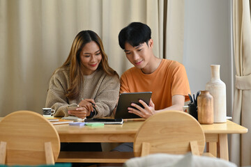 Fototapeta premium Happy Asian man and woman sitting on a table in a modern Livingroom looking at the tablet together. home and work concept