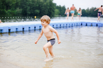 Little blond preschool boy having fun with splashing in a lake on summer day, outdoors. Happy child learning swimming. Active leisure with kids on vacations. Danger on domestic lakes