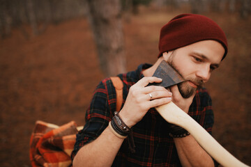 Bearded millennial man in plaid shirt is hiking with backpack, axe and firewood like lumberjack.