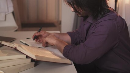 Study foreign language. Male student. Second degree. Busy man making notes from reference book sitting desk light shadow room interior.