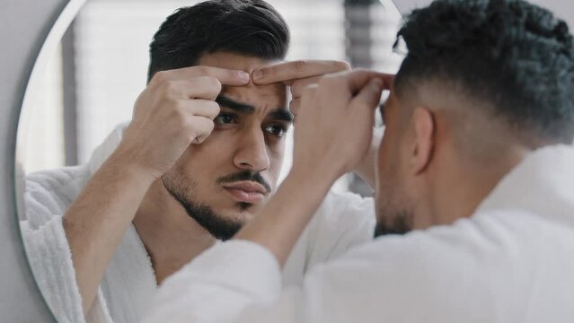 Unhappy arabian indian arab bearded man looking in bathroom mirror feeling worried about facial skin problem acne. Anxious millennial guy squeezing pimple touching face frustrated blackheads skincare