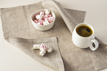 On a gray linen napkin, a white-pink marshmallow in a white bowl and a mug of coffee.