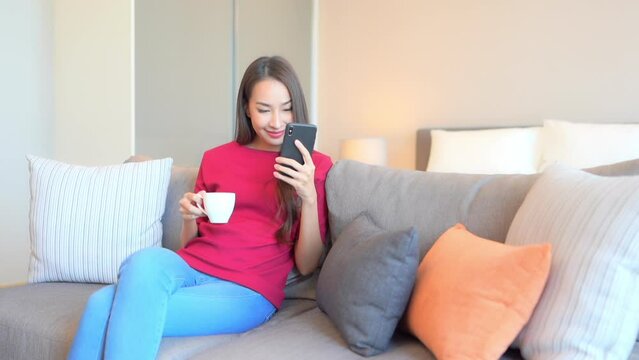 Sitting on the couch with a cup of coffee in one hand and a cell phone in the other, a pretty young woman reacts to her screen. 