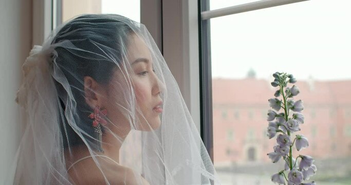 Sensual young asian woman seductively looks outside the window holding flower in her hand. Wedding photo session of charming sensual bride under white veil. Bridal day concept