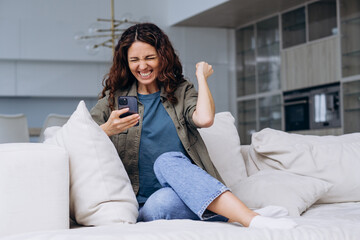Joyful curly woman with a phone in her hands raised her hand up from a very pleasant news at home sitting on the couch, waiting for a promotion at work or received a large order