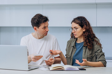 Fototapeta premium Husband and wife calculate the expense of finances sitting at home in the kitchen at the table and write it down in a notebook, a woman with checks in her hands makes excuses, a man gets upset