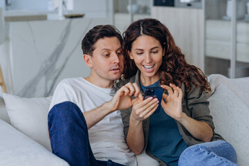 Excited married woman with long curly hair and brunet man smile happily reading good news in internet via smartphone sitting on sofa