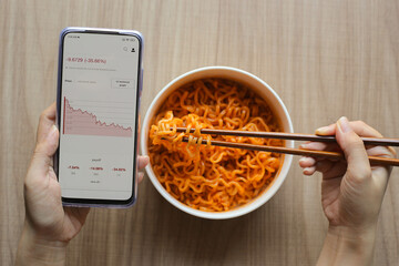 Use chopsticks eating noodles while using mobile phone monitor investment on stock, crypto trading which is continuously going down in red poor investor has no money to buy freshly cooked food