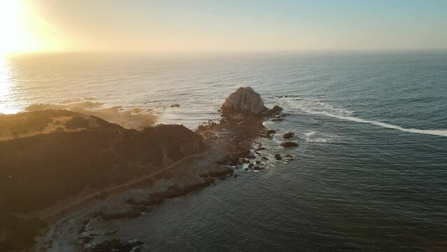 Aerial dolly in of ship sailing in sea near rocky shore in Pe&ntilde;a Blanca islet at golden hour, El Canelillo beach, Chile