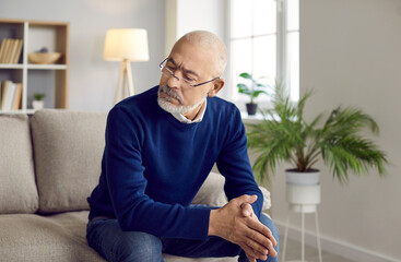 Candid shot of senior man deep in thought. Indoor portrait of bearded bald mature man in glasses sitting on sofa in living room, looking away at something off camera and thinking