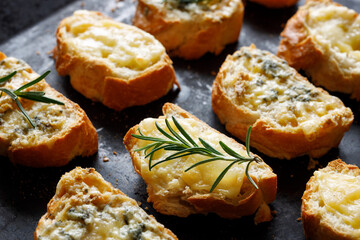 Crostini, cheese toasts  of sliced baguette with two types of cheese and fresh rosemary on a dark background, focus on the sandwich inside, close-up view.