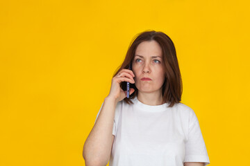 Portrait of a smiling european brunette woman wearing white T-shirt on yellow background talking on mobile phone.