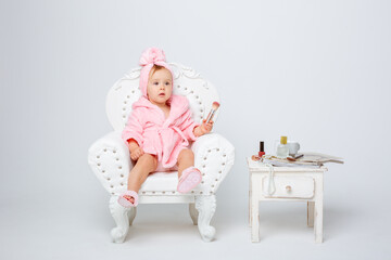 A cute little baby girl in a pink terry dressing gown with a chair and a mini dressing table with cosmetics is isolated on a white background, repeats the behavior of her mother