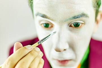 Close up of clown cosplayer. Mim is preparing makeup for performance. Man with green hair in burgundy suit. Theatre atmosphere. Male actor is covering his face by white greasepaint using brush.