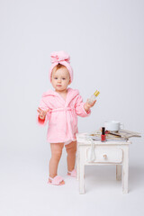 A cute little baby girl in a pink terry dressing gown with a mini dressing table and cosmetics is isolated on a white background, repeats the behavior of her mother