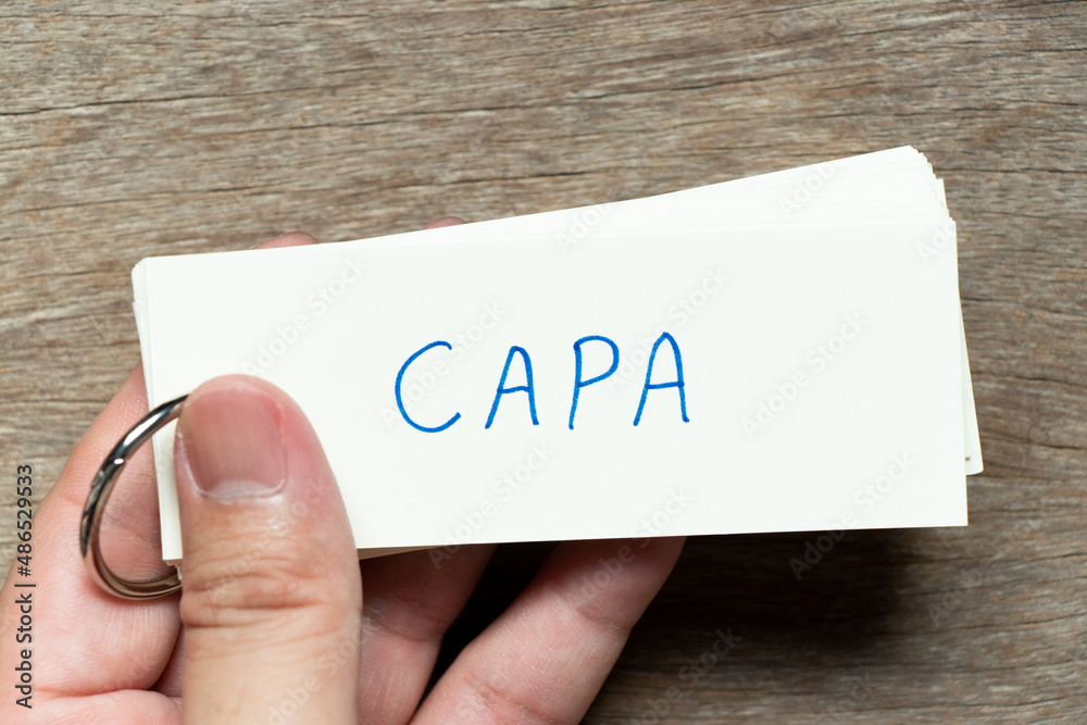 Poster Man hold the flash card with handwriting word CAPA (abbreviation of corrective action and preventive action) on wood background