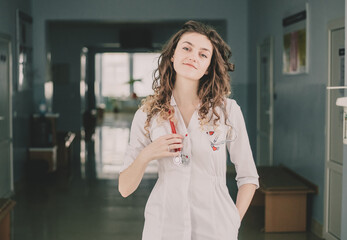 A woman doctor in a protective mask and a white coat stands against the backdrop of a polyclinic