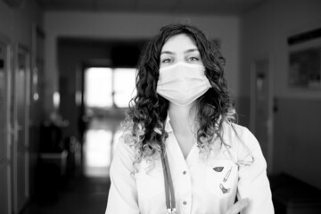 A woman doctor in a protective mask and a white coat stands against the backdrop of a hospital black and white photo
