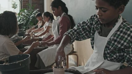 Titling up of cute Black boy sitting at pottery wheel and making craft of clay in workshop at daytime, female Caucasian teacher helping students on background