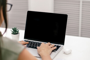 Fototapeta premium Young dark-haired businesswoman in a green dress and eyeglasses is typing on a laptop in a light office.Rear view, female hands and laptop screen close-up.Business concept.Copy space,selective focus
