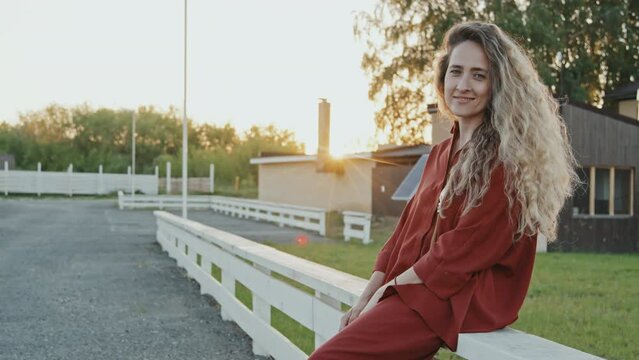 Medium long of blonde long-haired Caucasian woman wearing two-piece casual set, leaning on white fencing in neighborhood in summer evening, smiling and looking on camera