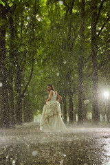 happy bride in a white wedding dress in the rain on an alley under big green trees walking peaying sitting and laughing