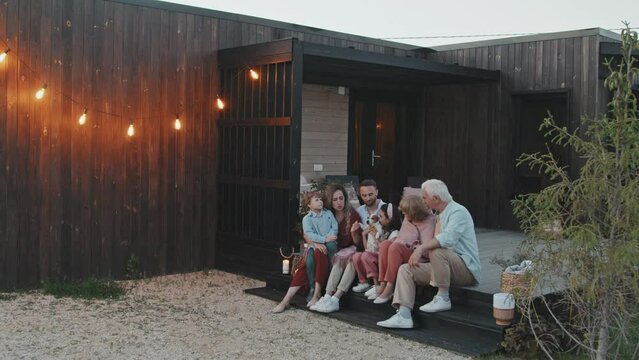Wide tracking in of grandparents, married Caucasian couple, two children and dog sitting on porch of summer house in summer evening, talking and smiling