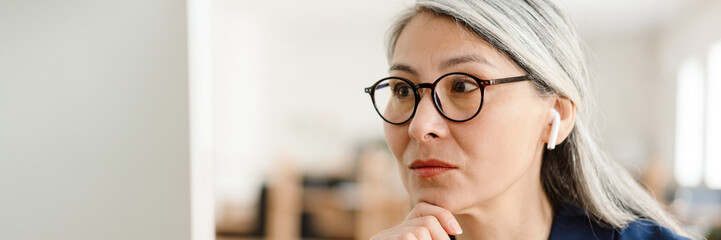 The portrait of an Asian woman in glasses and headphones looking at the computer monitor in the office