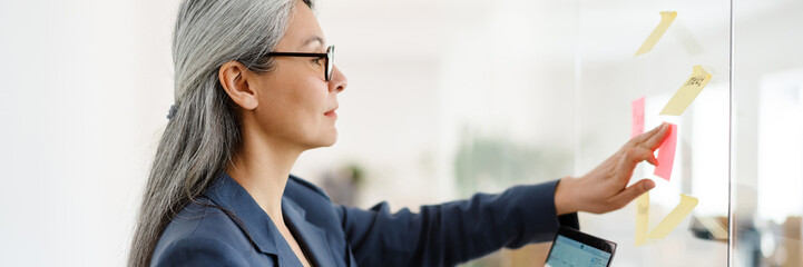 The side view of an Asian woman of adhesiving paper to the glass while holding a laptop in her hand in a the office