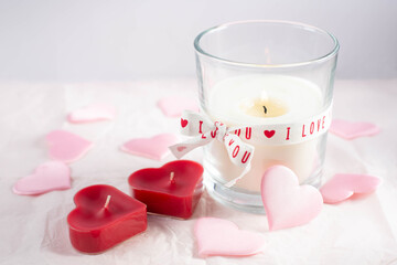 The theme is Valentine's Day. A burning candle and two red candles in the shape of hearts and decorative pink hearts are on a white background. Free space for text.