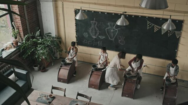 High angle of four multiethnic little pottery students wearing aprons, sitting in row at pottery wheels, making clay crafts in daylighted modern classroom, female teacher assisting