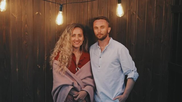 Medium portrait of blonde Caucasian woman and young blue-eyed man standing by wooden wall in light of incandescent lamps, looking and smiling on camera
