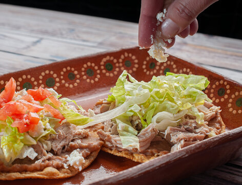 A hand putting cheese on top of tostadas on a clay or mud plate over a rusic table with chicken or carnitas, lettuce, tomato, onion, fried beans, spicy red salsa and cream.