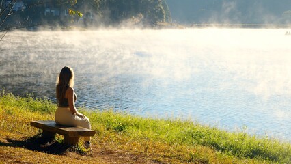 Travel woman relax on nature near misty lake. Female traveler sitting on bench, enjoy fog morning on river. Calmness, relaxation, meditation, freedom, beauty of nature concept