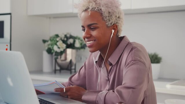 Young African American woman cinephile in wired headphones watching video podcast or movie in laptop relaxing during lunch break smiling holding notepad and pencil sits at table in home kitchen.